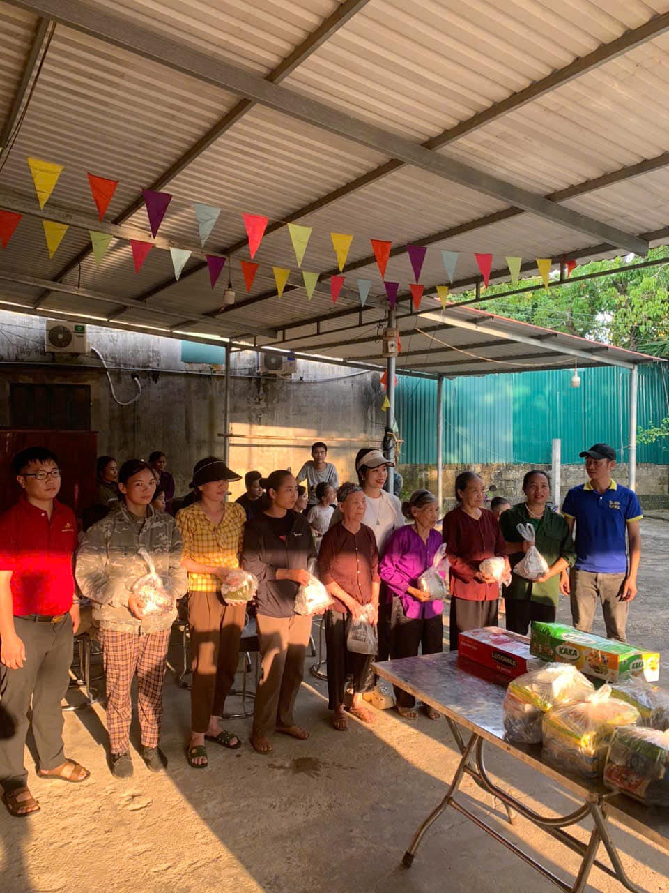 Nhóm người cao tuổi và thanh niên nhận quà từ thiện dưới mái hiên trang trí cờ nhiều màu