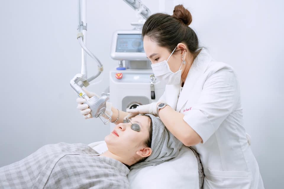 Bác sĩ da liễu đeo khẩu trang dùng máy laser điều trị da mặt cho khách hàng nằm trên giường spa